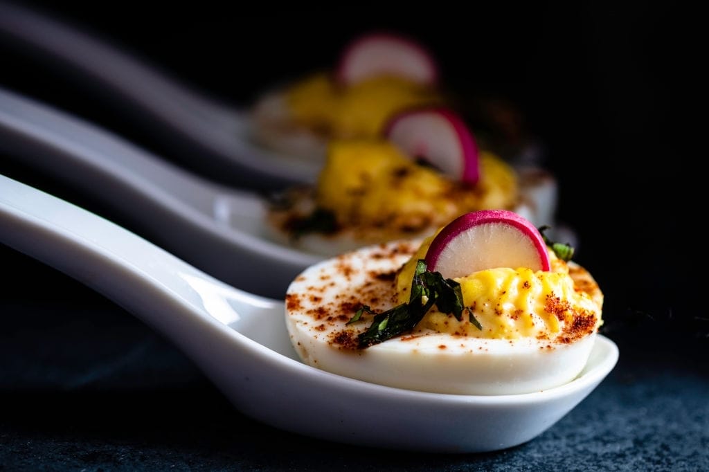 Deviled eggs garnished with smoked spanish paprika, sliced radishes, and chopped fresh basil; served on individual ceramic spoons.